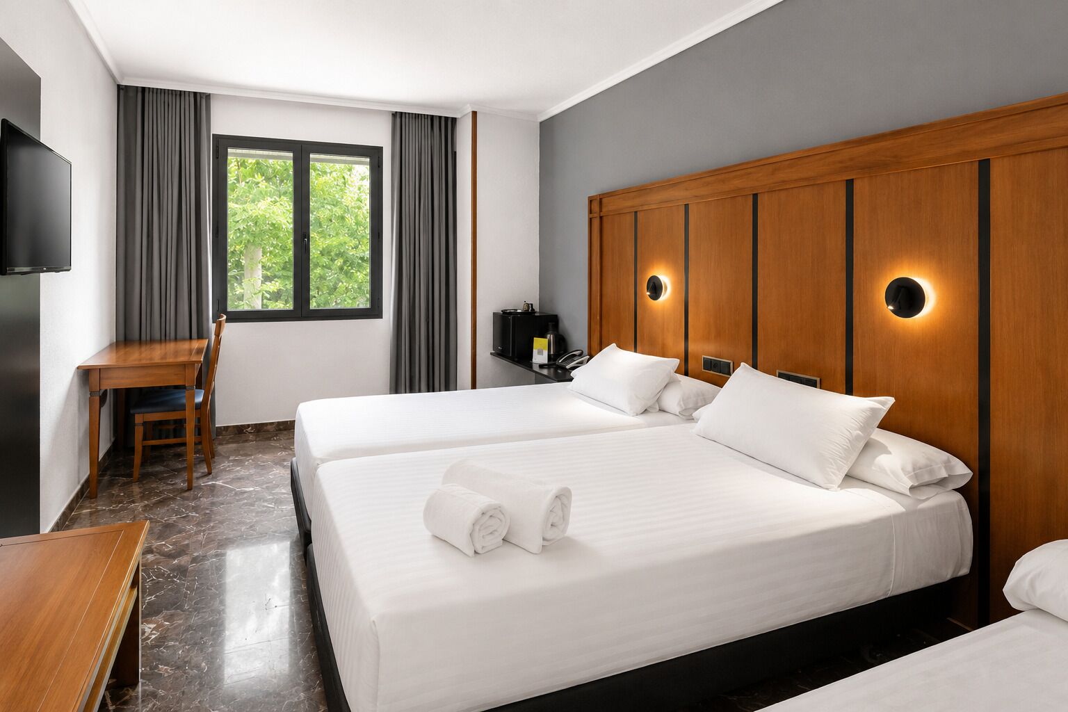 A hotel room with two beds, a desk, a TV, and a window with curtains.