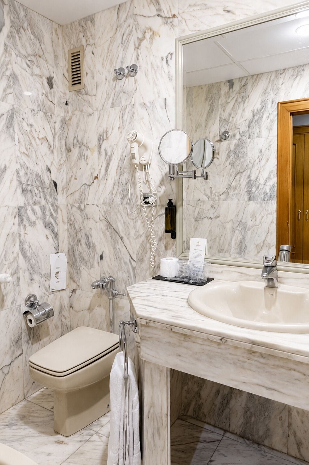 Une salle de bain avec un mur en marbre, un lavabo, des toilettes, un porte-serviettes, un miroir et une douche.