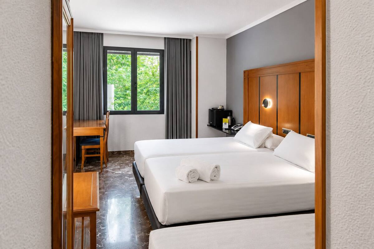 Una habitación de hotel con cama, escritorio, silla, mesita de noche y ventana con cortinas.
