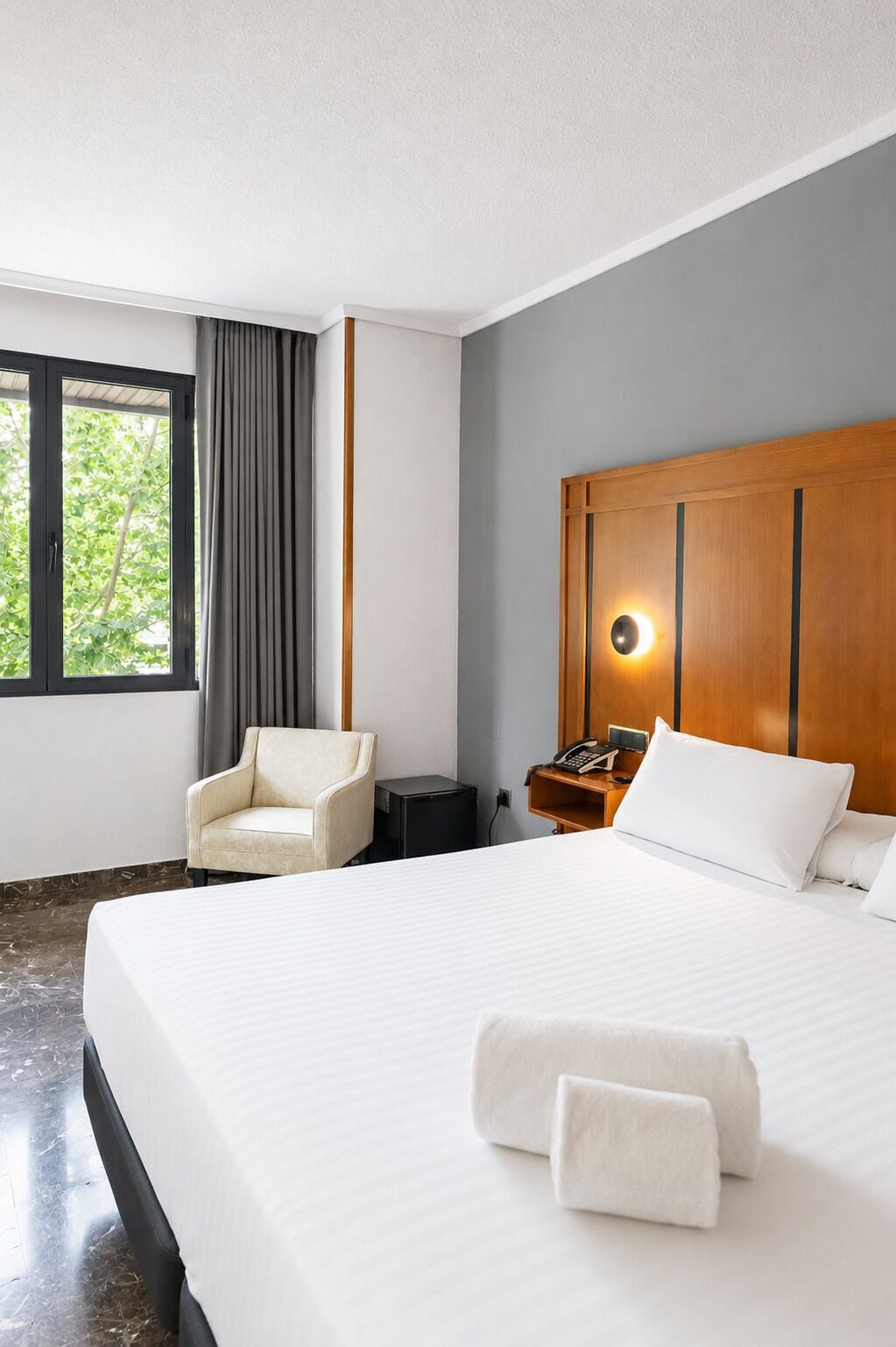 Una camera d'albergo con un letto grande, una poltrona, un comodino con il telefono e una finestra con tende.
