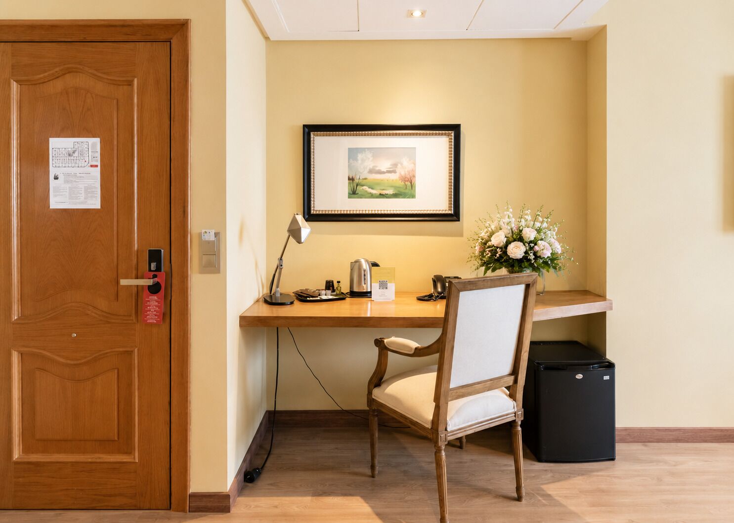 A hotel room with a wooden desk, a chair, a framed picture on the wall, a vase with flowers, and a black trash bin.
