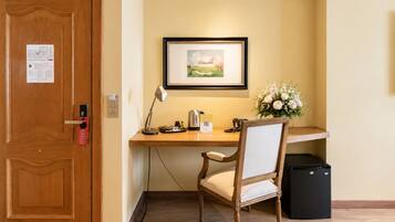 A hotel room with a wooden desk, a chair, a framed picture on the wall, a vase with flowers, and a black trash bin.