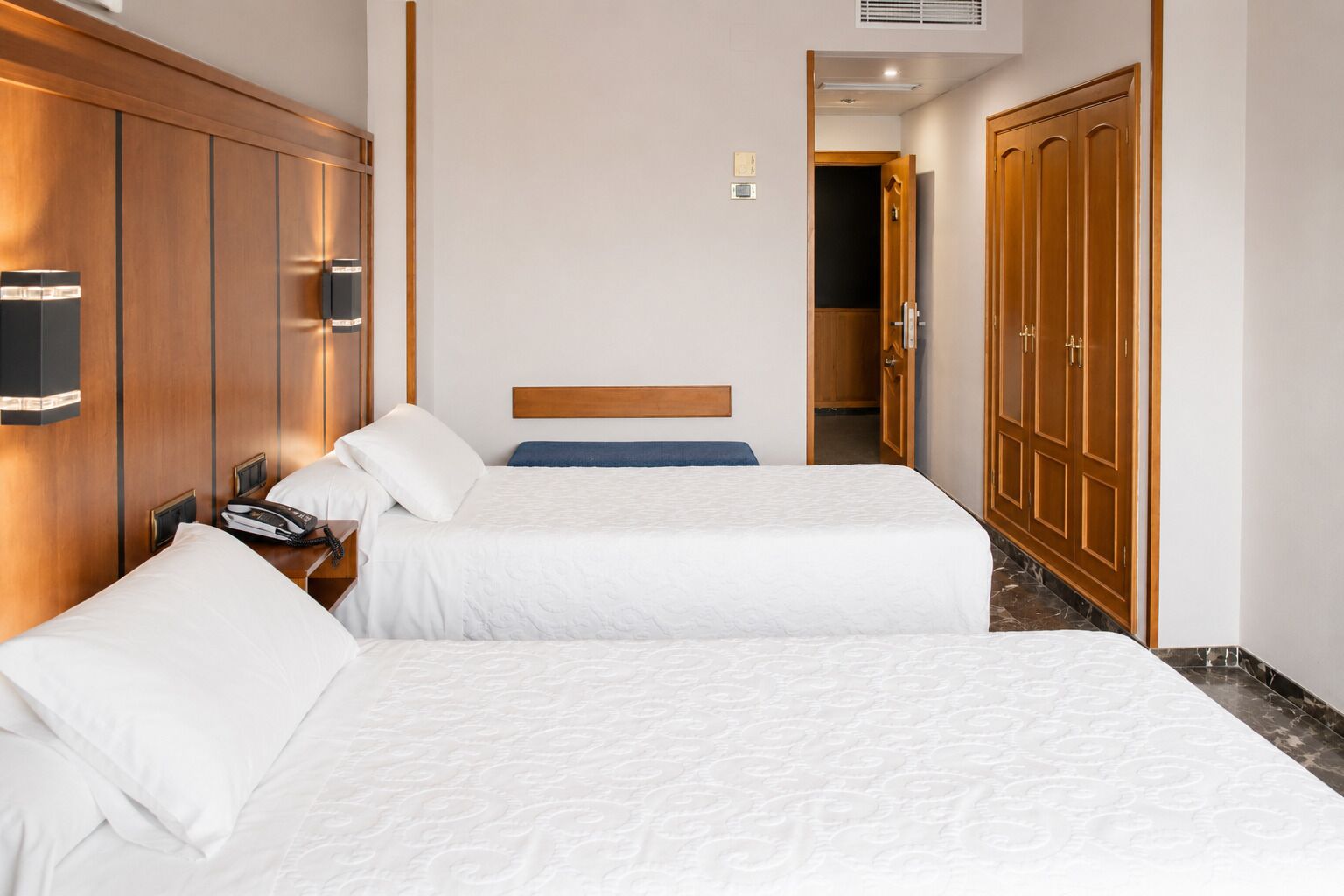 A hotel room with two beds, wooden wardrobe, and a small desk with a telephone.