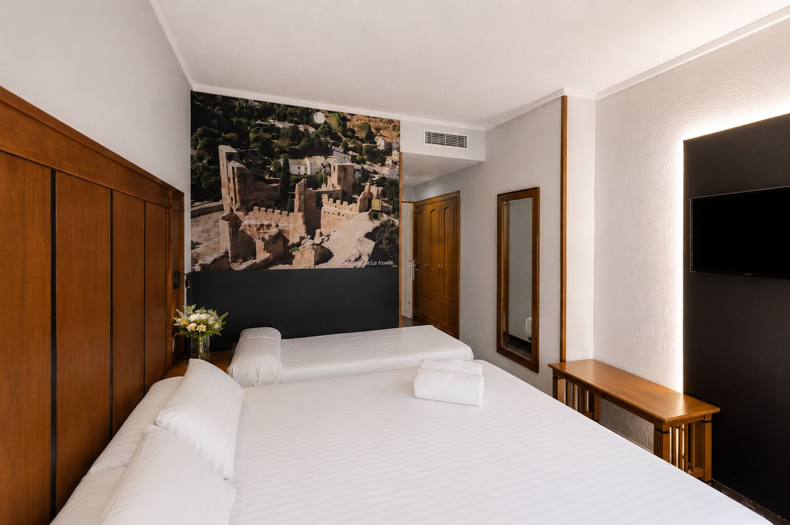 Een hotelkamer met een groot bed, een houten garderobe, een spiegel, een flatscreen televisie en een muurschildering.