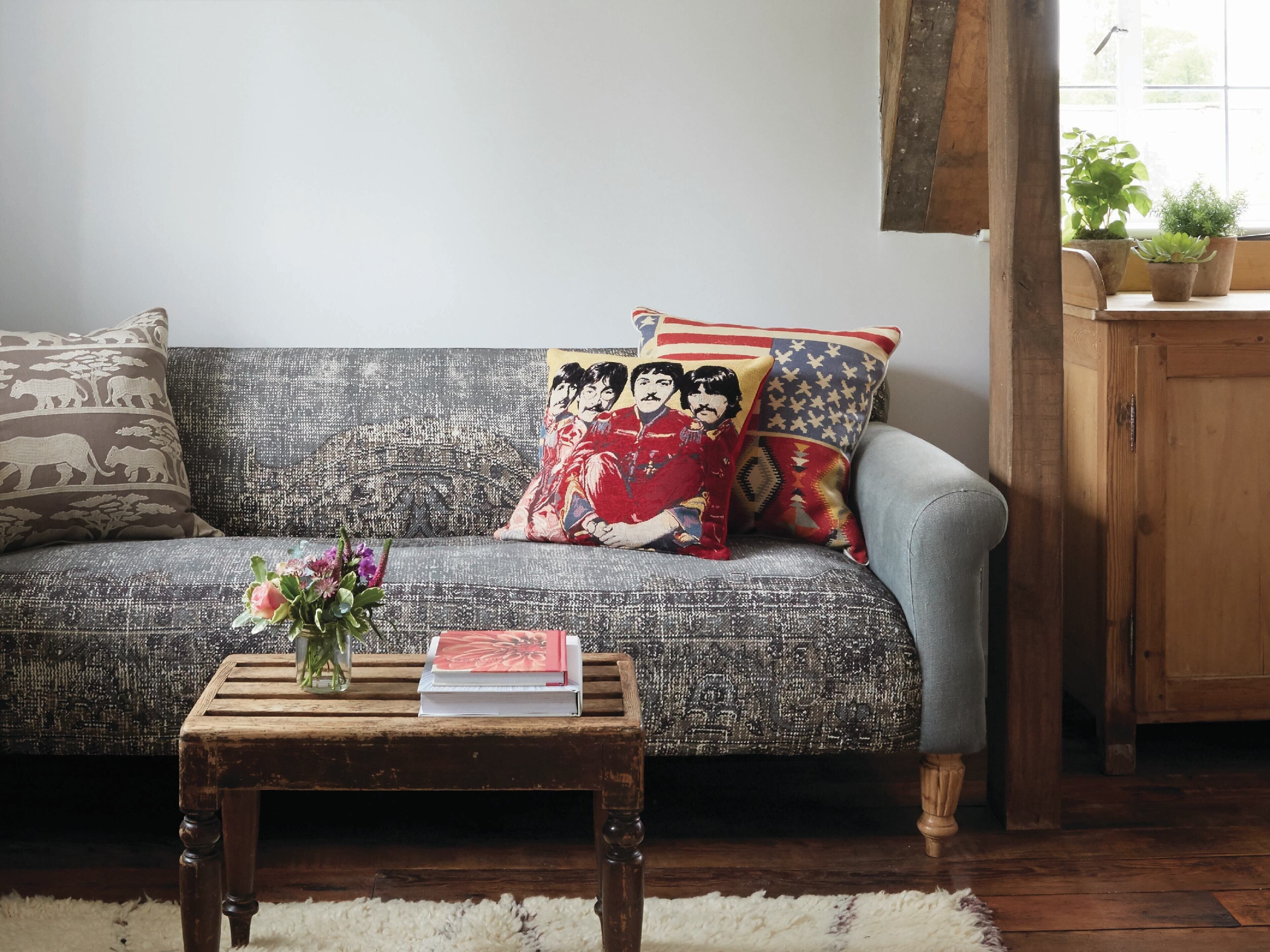 Un divano con cuscini decorati, un tavolino di legno con un libro e dei fiori, e una finestra con piante.