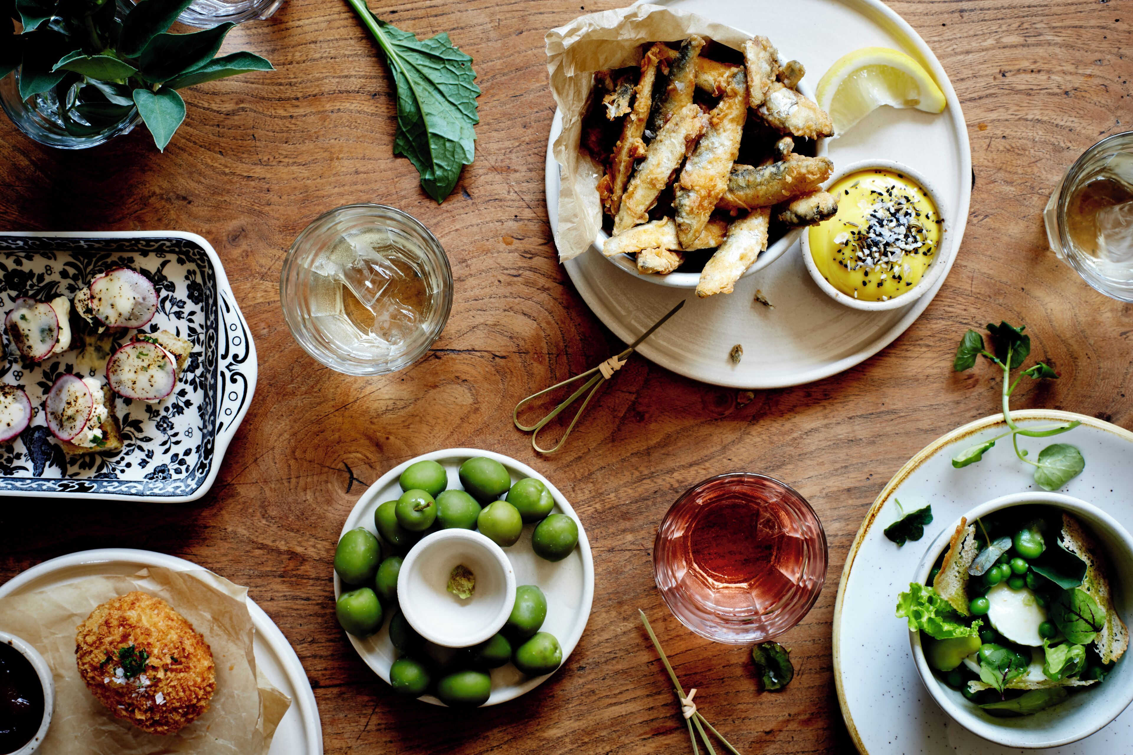Diverse gerechten op een houten tafel, waaronder gefrituurde snacks, een schaal olijven en een glas water.