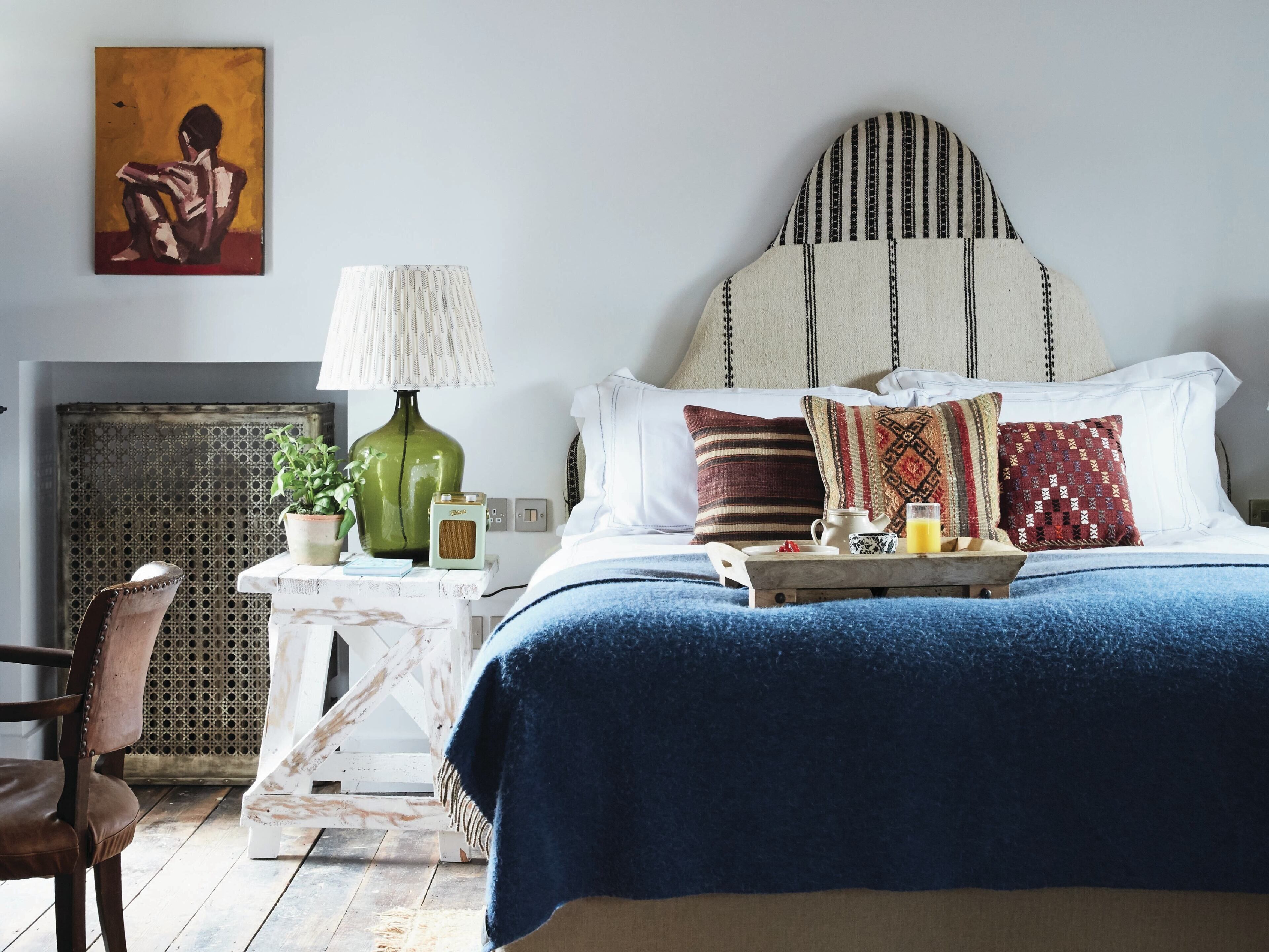 A bedroom with a bed, pillows, a bedside table with a lamp, a chair, and a painting on the wall.