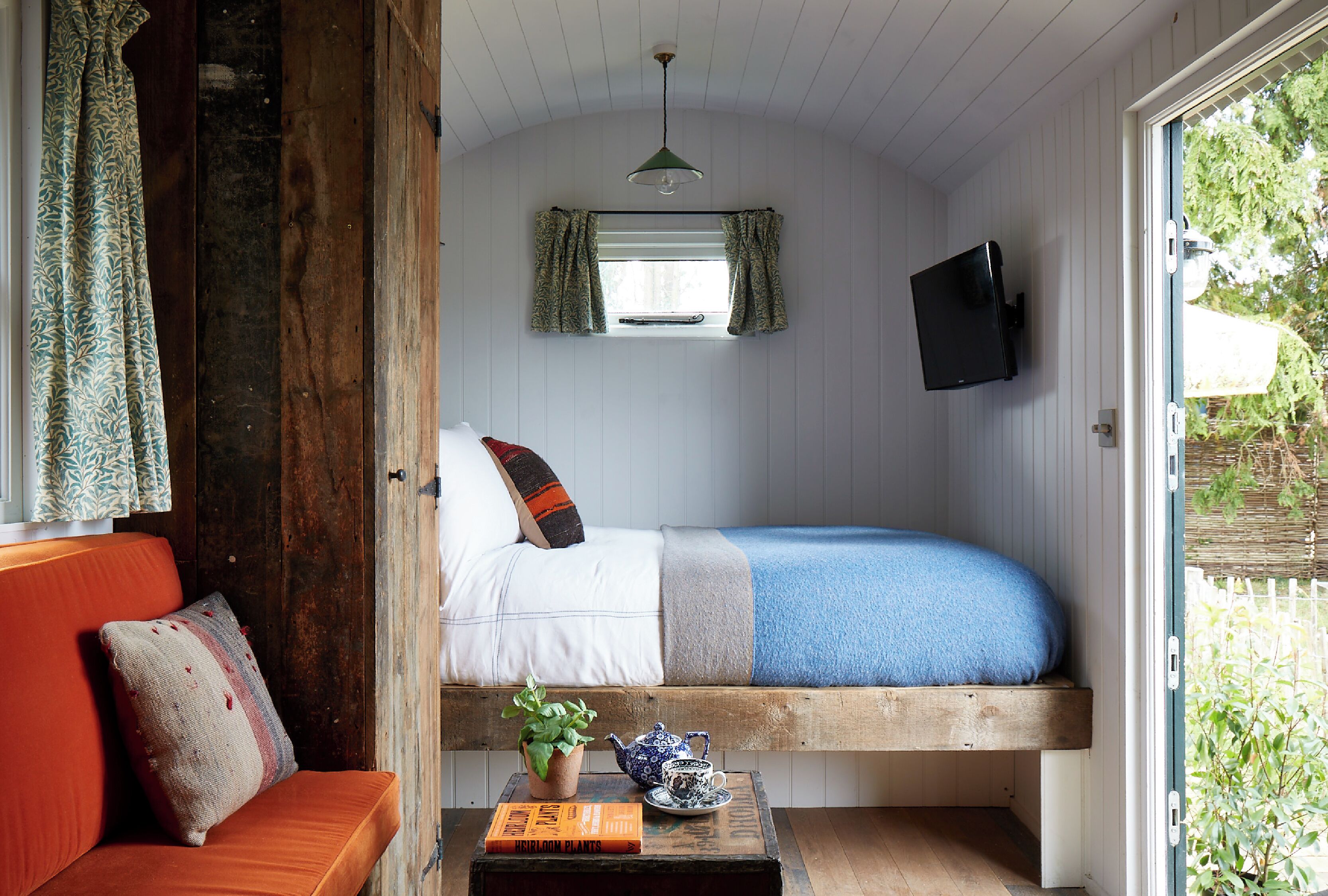 Una habitación acogedora con una cama, un sofá, una mesita con un libro, una planta en maceta y un televisor.