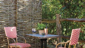 Un área de estar al aire libre rústica con un muro tejido, dos sillas de mimbre con cojines estampados y una mesa con una botella y vasos.
