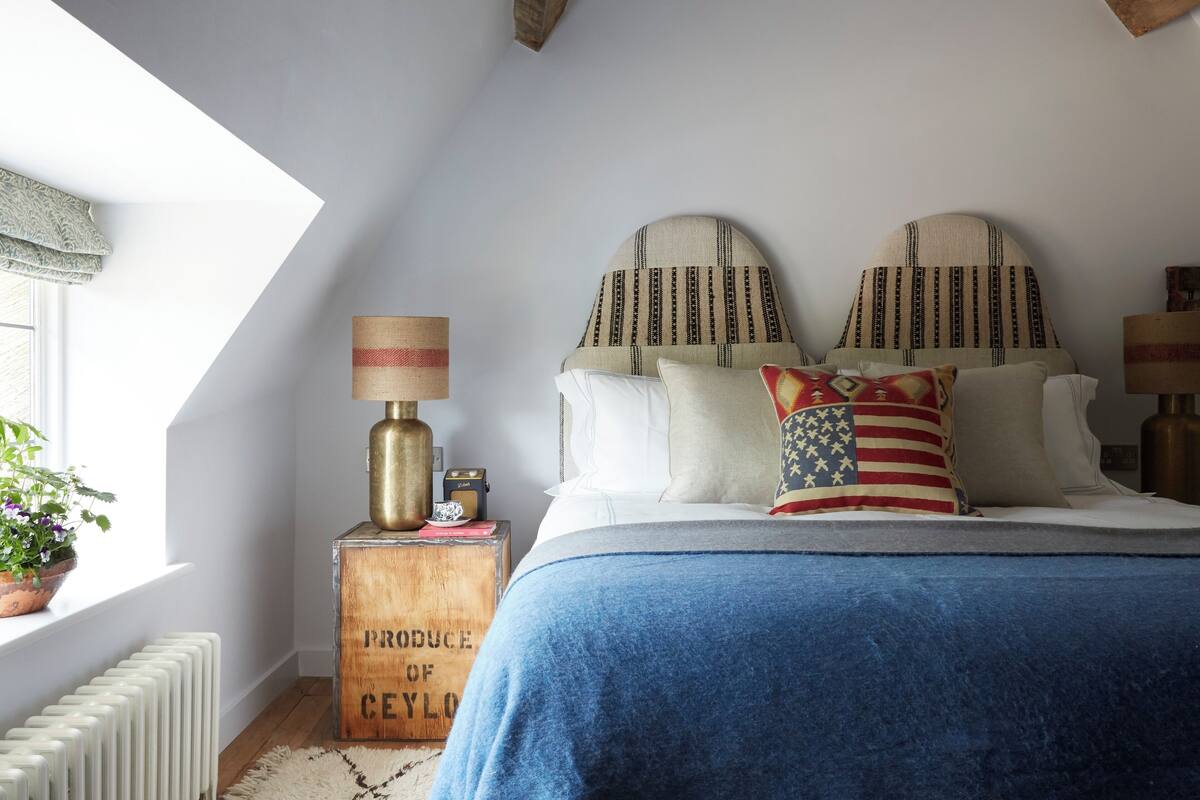 A bedroom with a sloped ceiling, a bed with a blue blanket, patterned pillows, a lamp, and a radiator.