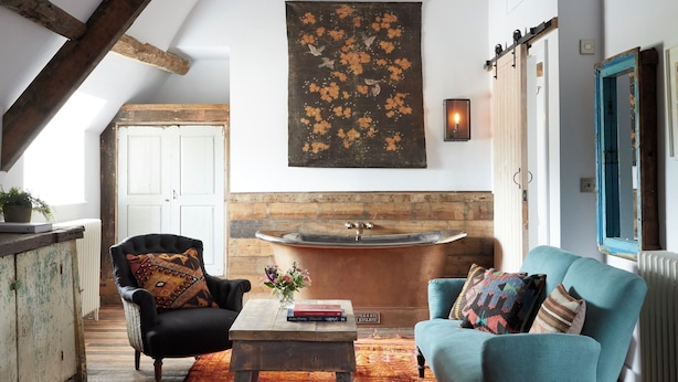 Une pièce de style « loft », avec des poutres en bois apparentes, une grande fenêtre, une baignoire rustique et un fauteuil bleu.