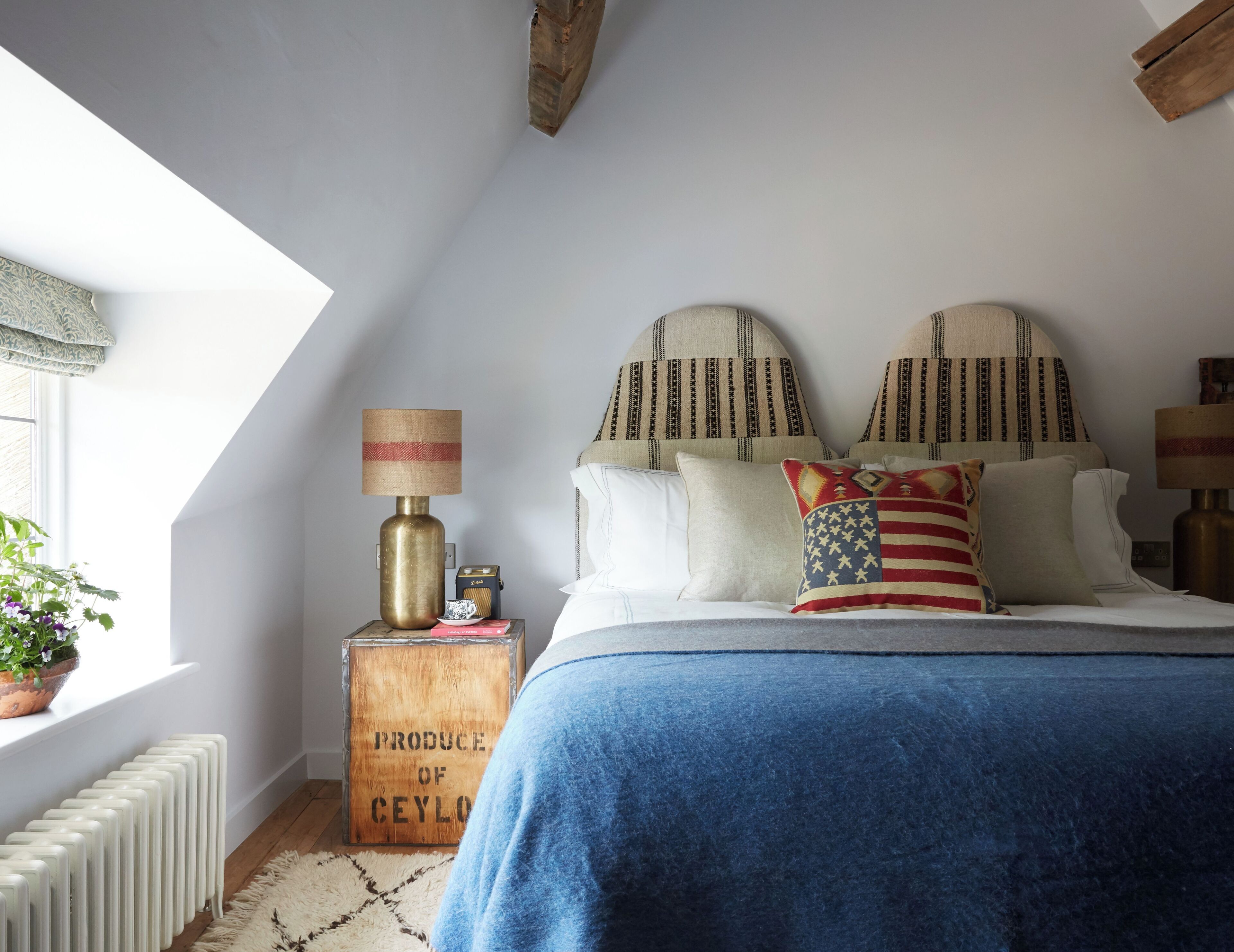 A bedroom with a sloped ceiling, a bed with a blue blanket, two decorative pillows, a lamp, a vintage-style radiator, and a window with a plant.