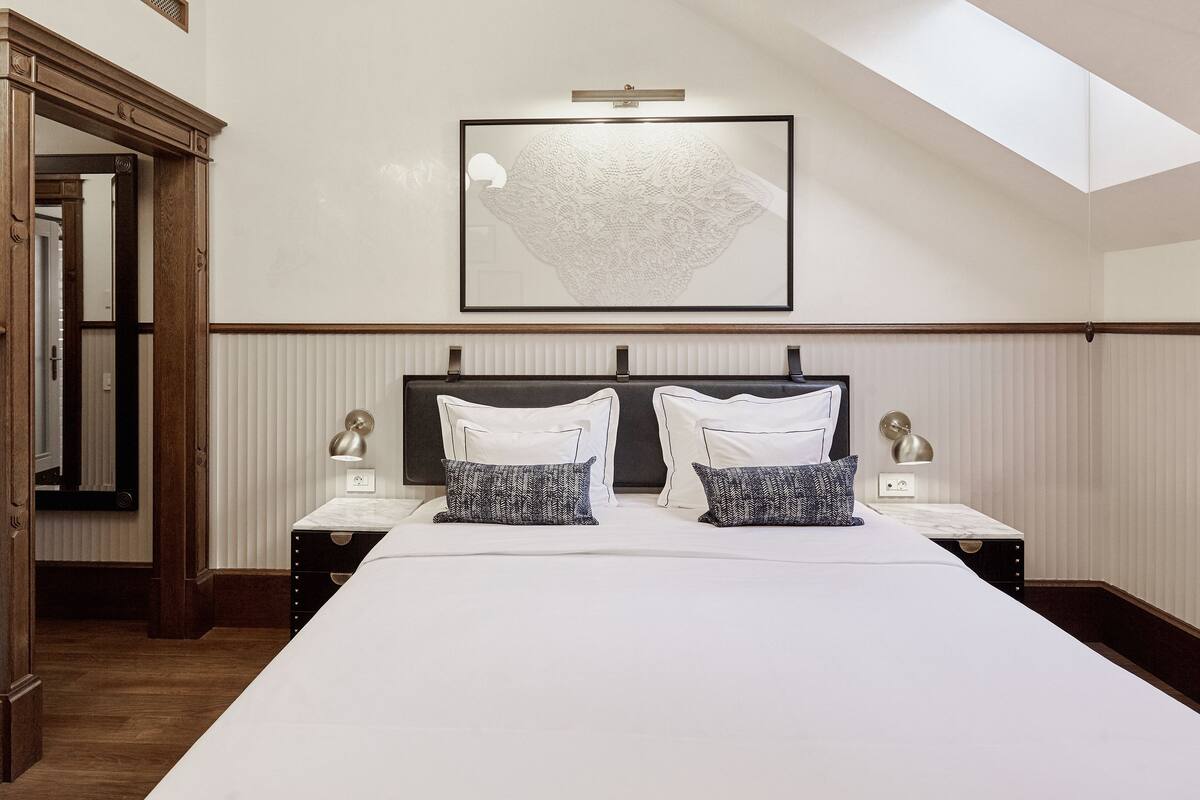 A hotel room with a large bed, two bedside tables, a mirror, a framed artwork, and a skylight.