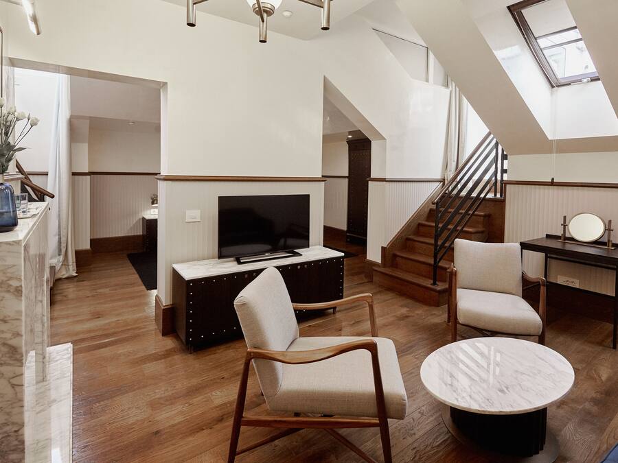 A spacious living area with a wooden floor, a round marble coffee table, a television on a dark wooden stand, and a staircase leading to an upper level.