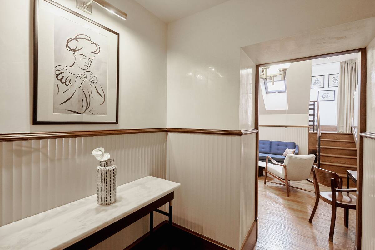Un pasillo con un banco, una obra de arte enmarcada, un jarrón y una escalera que conduce a una sala de estar con sofá y sillones.