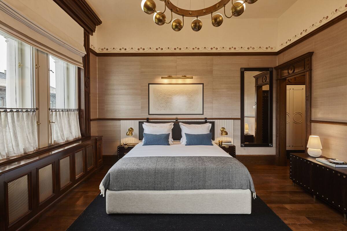 A large bed with a headboard, a bedside table with a lamp, a large mirror, and a window with curtains.