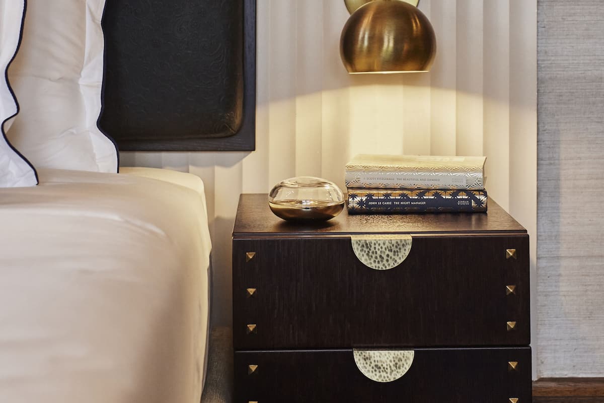 A neatly made bed with a headboard, a bedside lamp, a stack of books, and a small decorative item on a wooden nightstand.