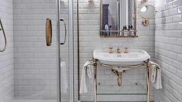 Un baño con ducha envuelta en vidrio, un lavamanos blanco con detalles dorados y un espejo con marco dorado.