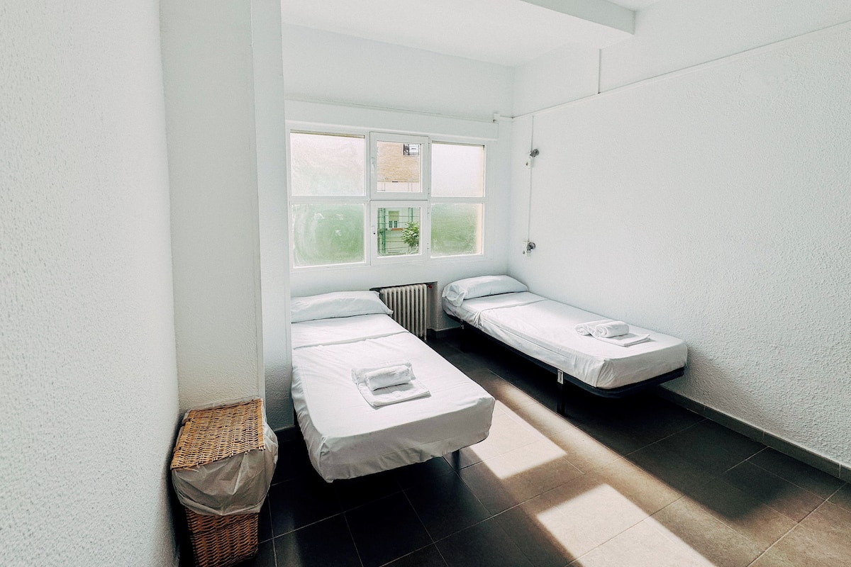 Quarto pequeno e luminoso com duas camas individuais, uma janela e um radiador.