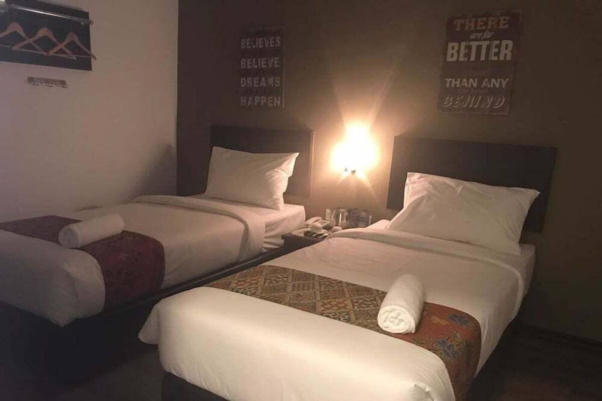 Deux lits jumeaux dans une chambre d’hôtel, avec un lampadaire fixé au mur et des affiches inspirantes.