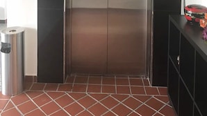Ein moderner Aufzug mit digitaler Steuerung, einem Mülleimer und einer kleinen Ablagefläche mit Gegenständen rechts.