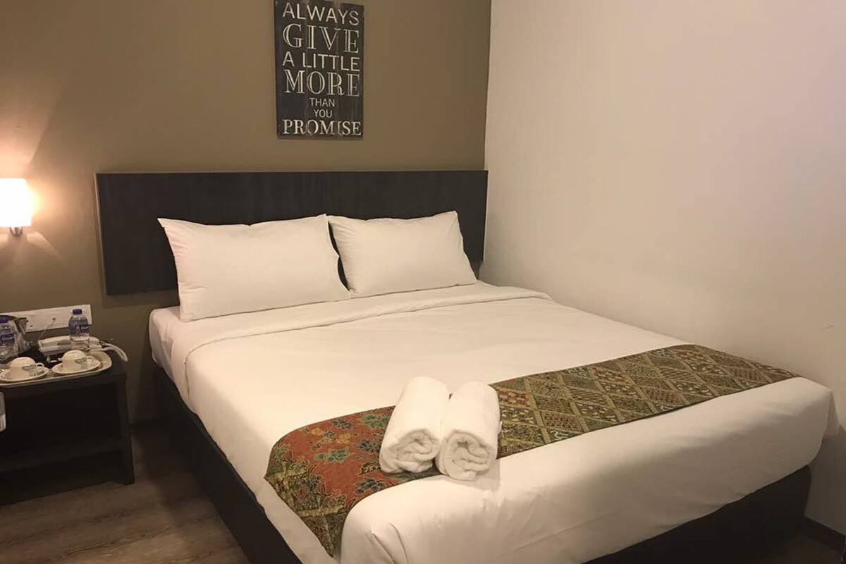 Un lit bien fait, avec du linge de lit blanc et un couvre-lit à motifs, une table de chevet avec une lampe et un téléphone, et une citation inspirante accrochée au mur.