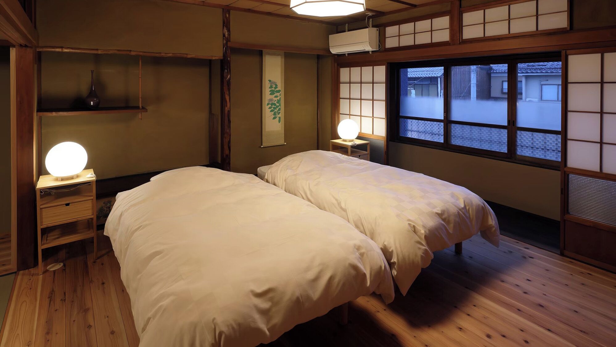 Une pièce de style japonais traditionnel, avec deux lits, un plancher en bois, une petite table de chevet avec une lampe et une fenêtre dotée de panneaux coulissants en papier.