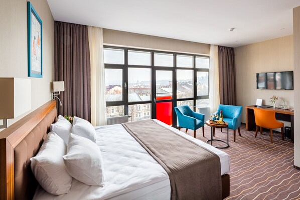 Habitación de hotel con una cama grande, dos sillas, un escritorio, un televisor y una ventana con vistas a la ciudad.