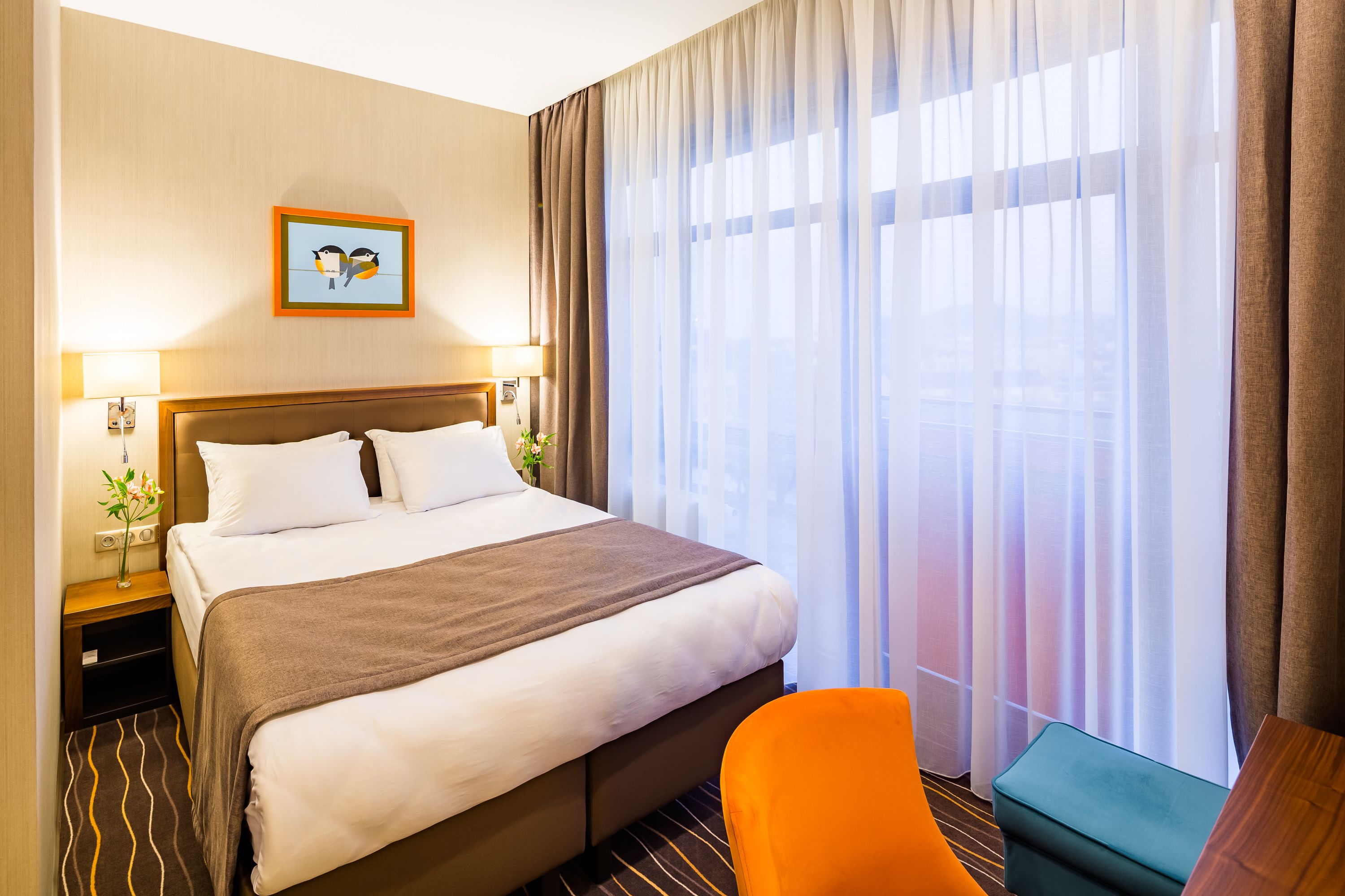 A hotel room with a large bed, a nightstand with a lamp, a small table with a vase of flowers, and a view of the city through sheer curtains.
