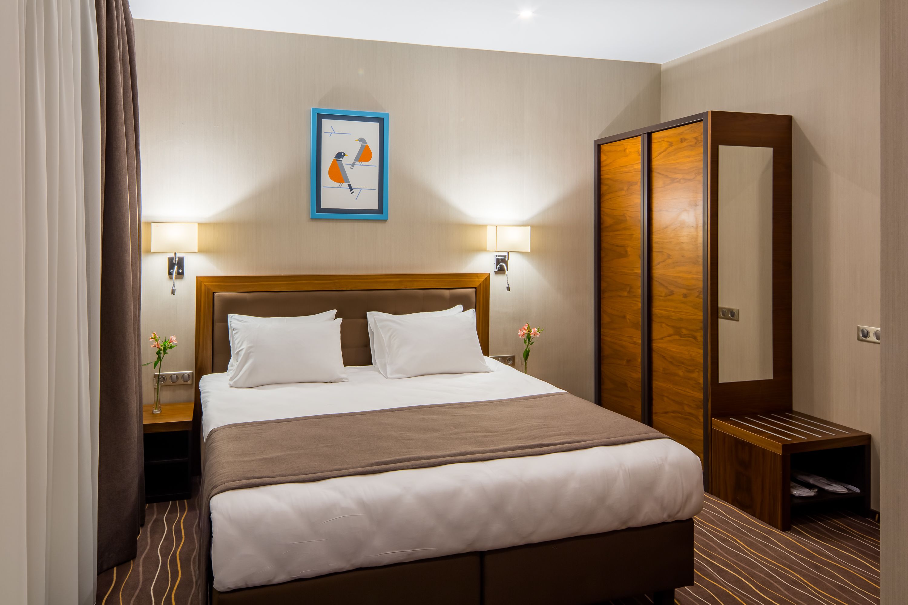A hotel room with a large bed, two bedside lamps, a wooden wardrobe, and a framed picture on the wall.