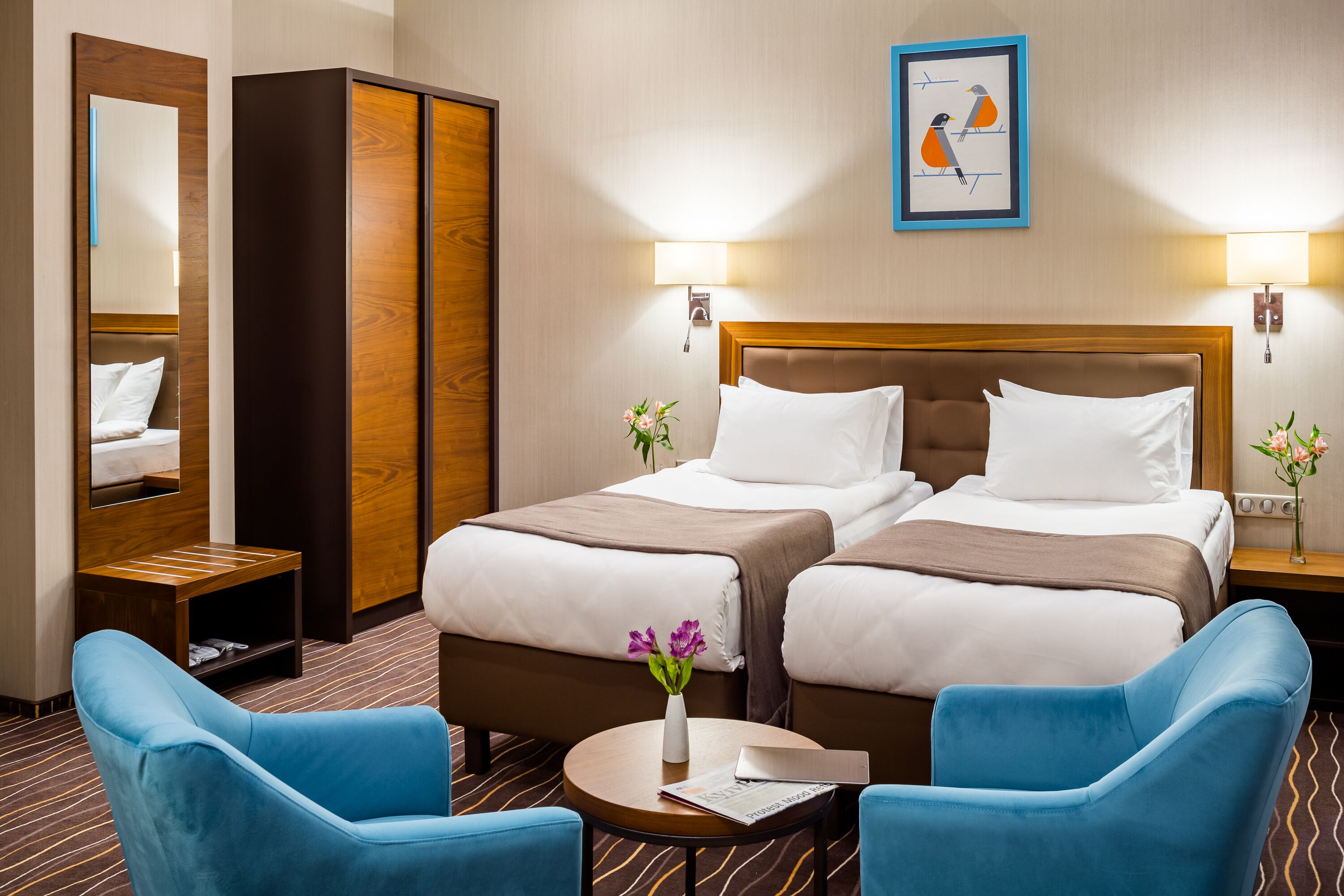 A hotel room with two beds, a mirror, a wardrobe, a desk, and two blue chairs.