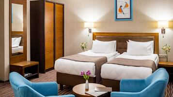 A hotel room with two beds, a mirror, a wardrobe, a desk, and two blue chairs.