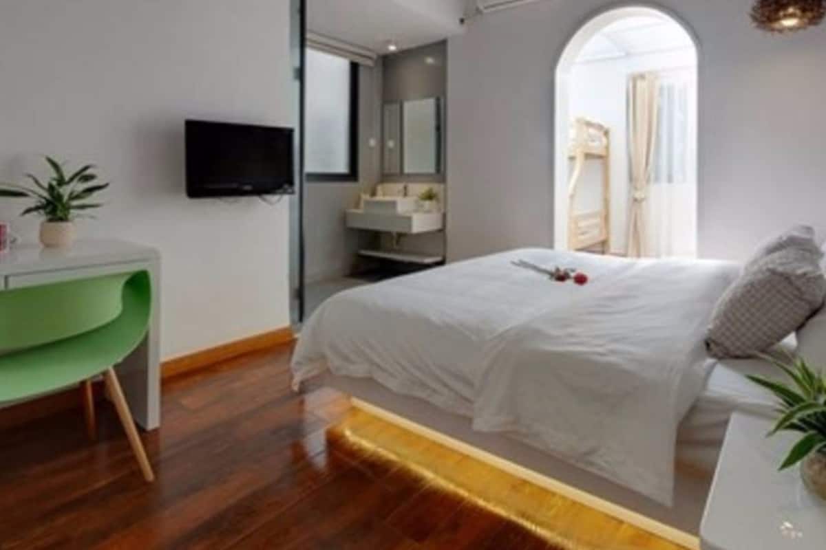 A modern bedroom with a wooden floor, a green chair, a bed with white bedding, a TV on the wall, and a bathroom visible through an archway.