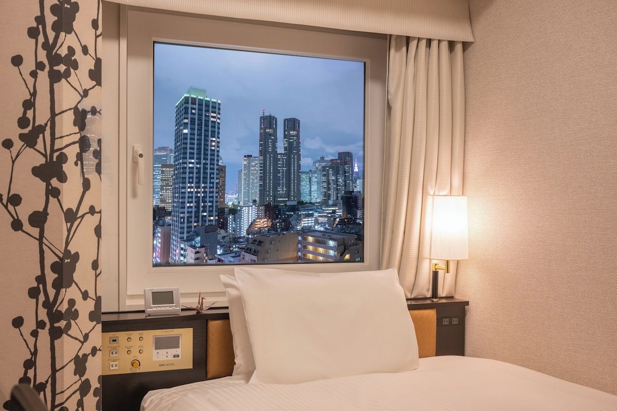 A hotel room with a large window offering a cityscape view, a bed with white linens, a bedside table with a lamp, and a control panel with room amenities.
