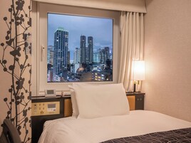 Habitación de hotel con un ventanal grande que ofrece vistas a la ciudad, una cama con sábanas blancas, una mesita de noche con lámpara y un panel de control con servicios de la habitación.