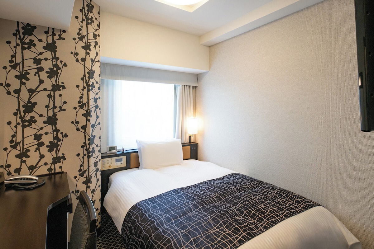 A hotel room with a bed, a desk, a telephone, a nightstand, and a window with curtains.