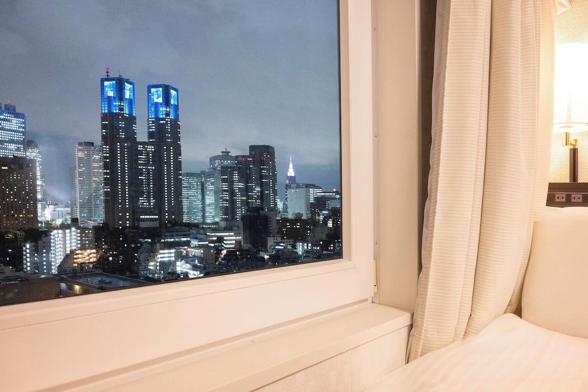 A hotel room with a cityscape view through a window, a bed, and a bedside lamp.