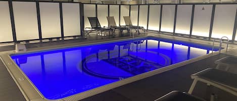 An indoor swimming pool with a blue-lit water feature, surrounded by lounge chairs and a wooden deck.