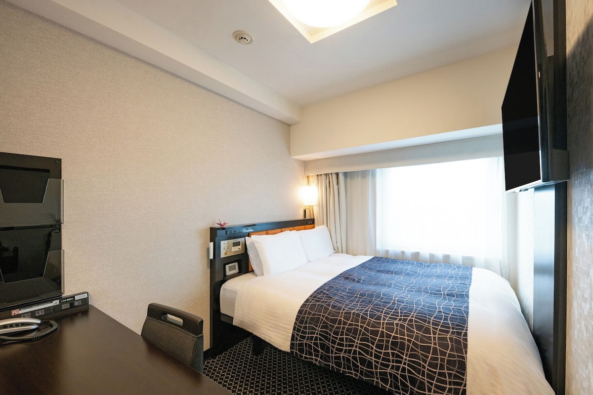 A hotel room with a bed, a desk with a television, a telephone, and a window with curtains.