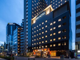 Un hotel moderno con un llamativo letrero de 'APA HOTEL', varios pisos y una entrada bien iluminada.