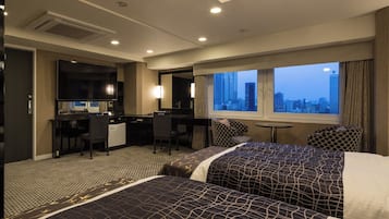 A hotel room with two beds, a desk, a chair, a small table, and a view of the cityscape.