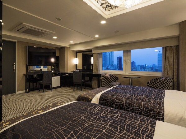 Habitación de hotel con dos camas, un escritorio, una silla, una mesita y vistas a la ciudad.