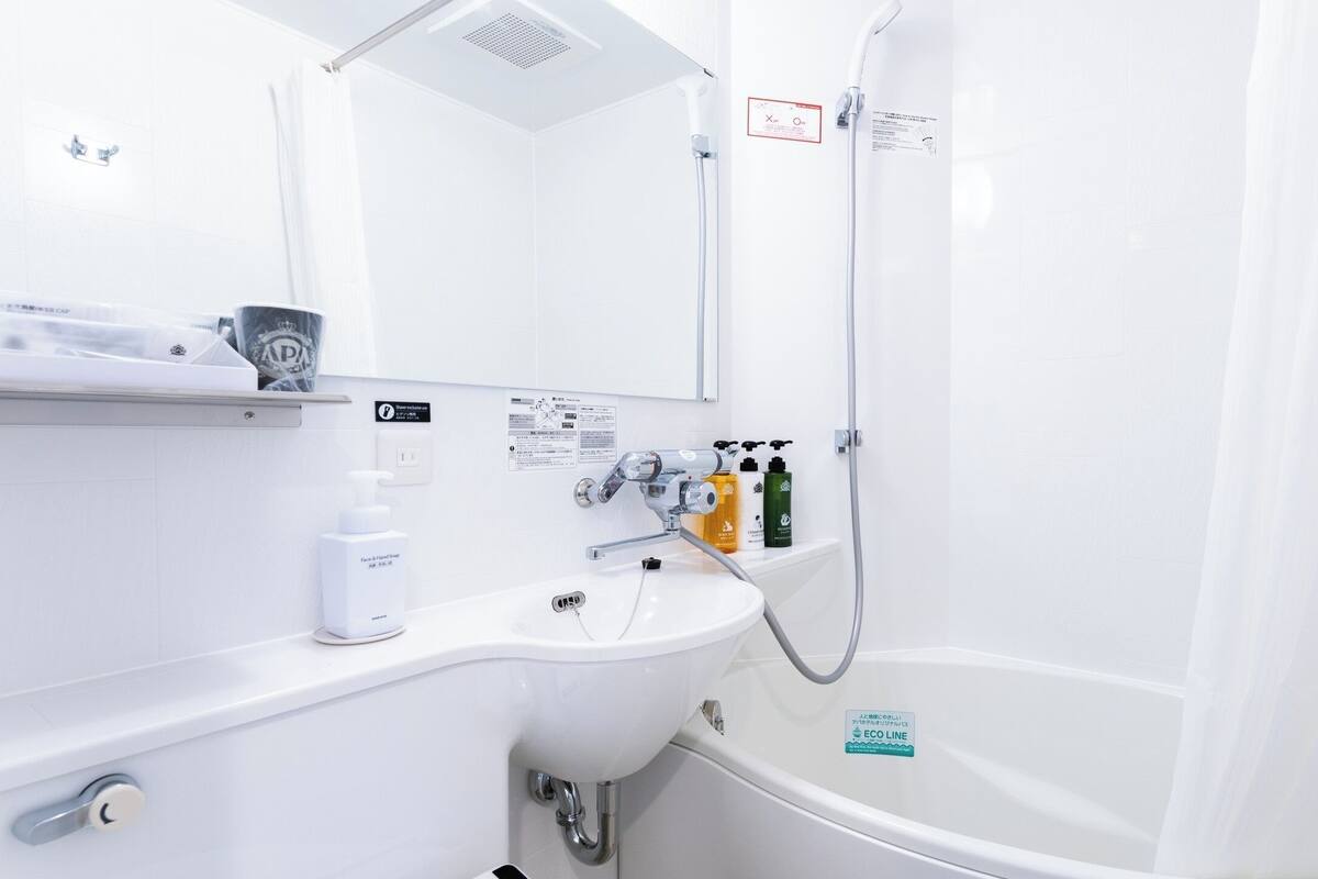 A modern bathroom with a white sink, a mirror, a shower, and a bathtub. There are toiletries on the sink and a sign indicating the bathroom's eco-friendly features.