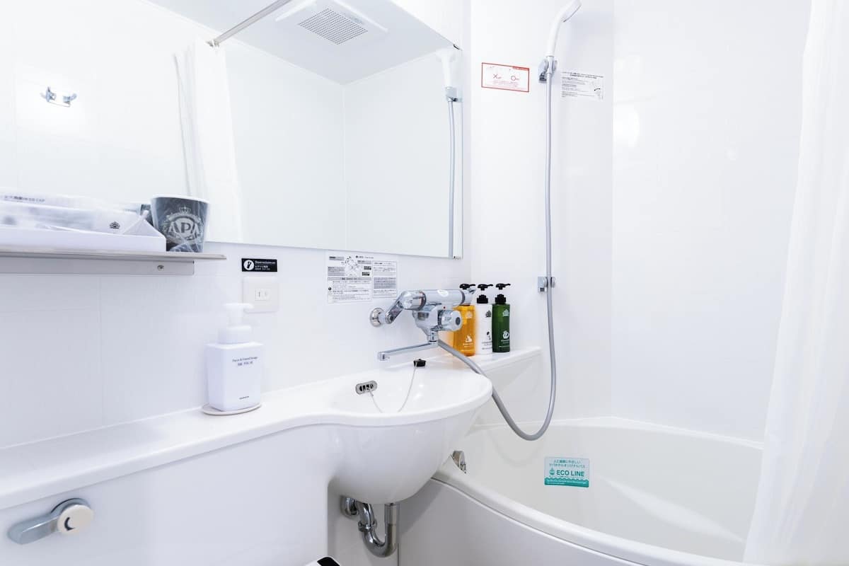 A modern bathroom with a white sink, a mirror, a shower, and a bathtub. There are toiletries on the sink and a sign indicating the bathroom's eco-friendly features.