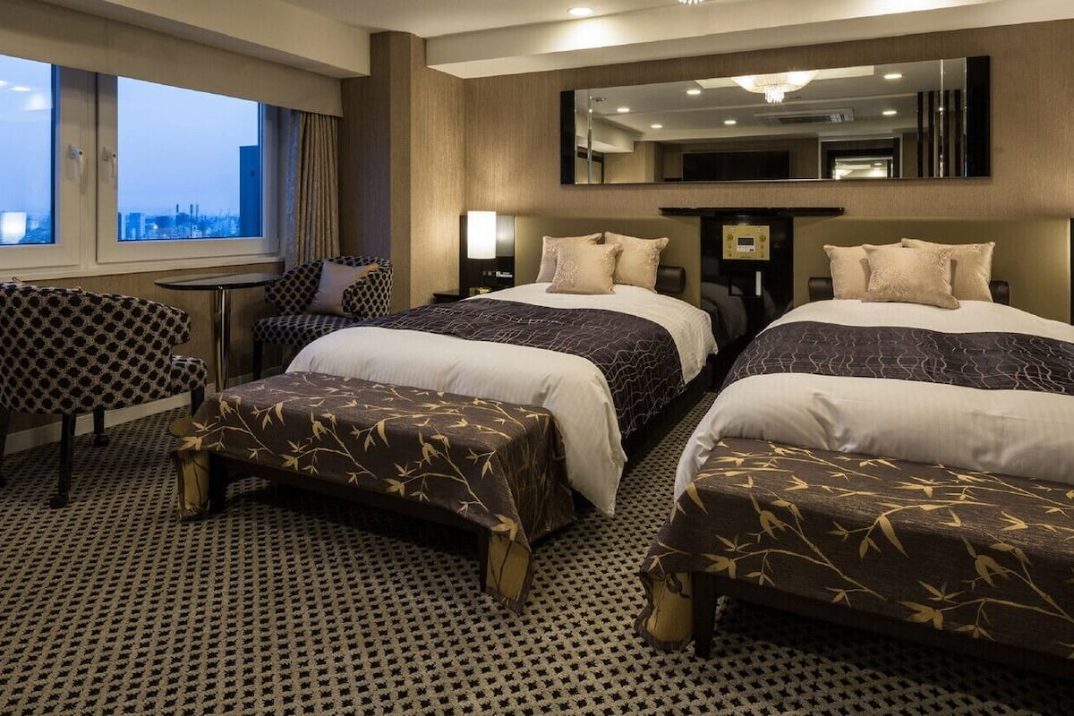 Habitación de hotel con dos camas, un escritorio, una silla y un ventanal con vistas a la ciudad.