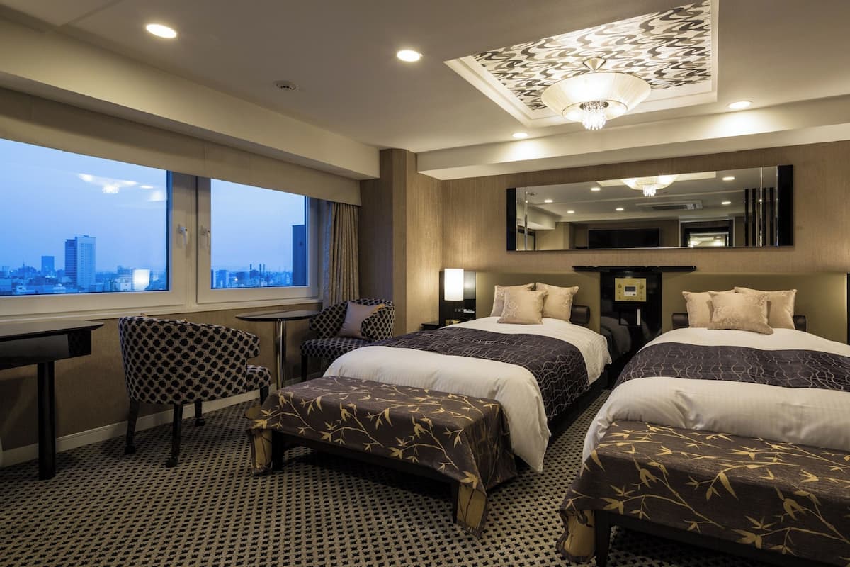A hotel room with two beds, a desk, a chair, a large mirror, and a view of the cityscape.