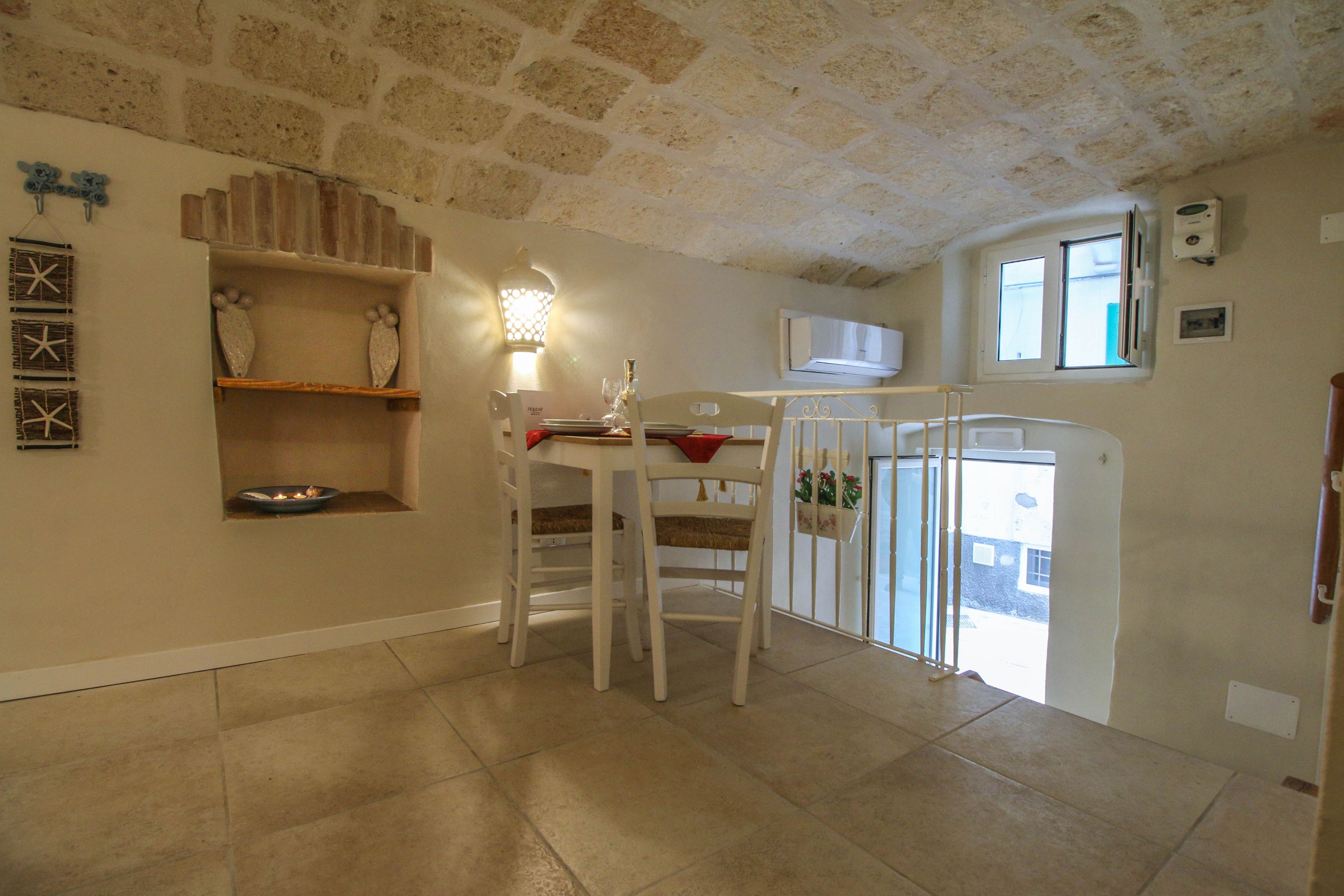 A dining area with a table and chairs, a wall-mounted shelf with decorative items, a wall-mounted light fixture, and a window with an air conditioning unit.