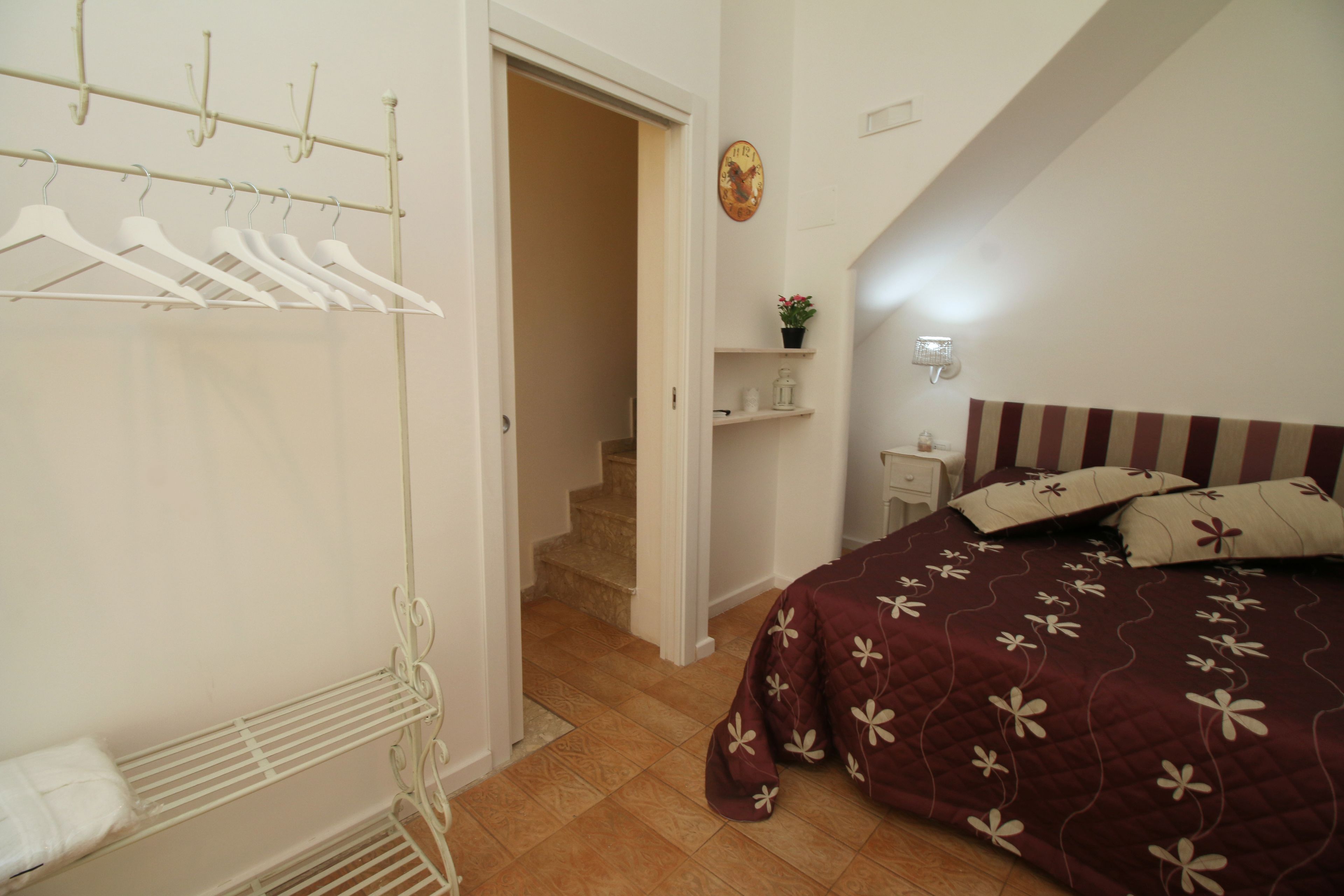 Un dormitorio con cama, un armario con ropa colgada, una mesita de noche pequeña y un estante en la pared con una planta.