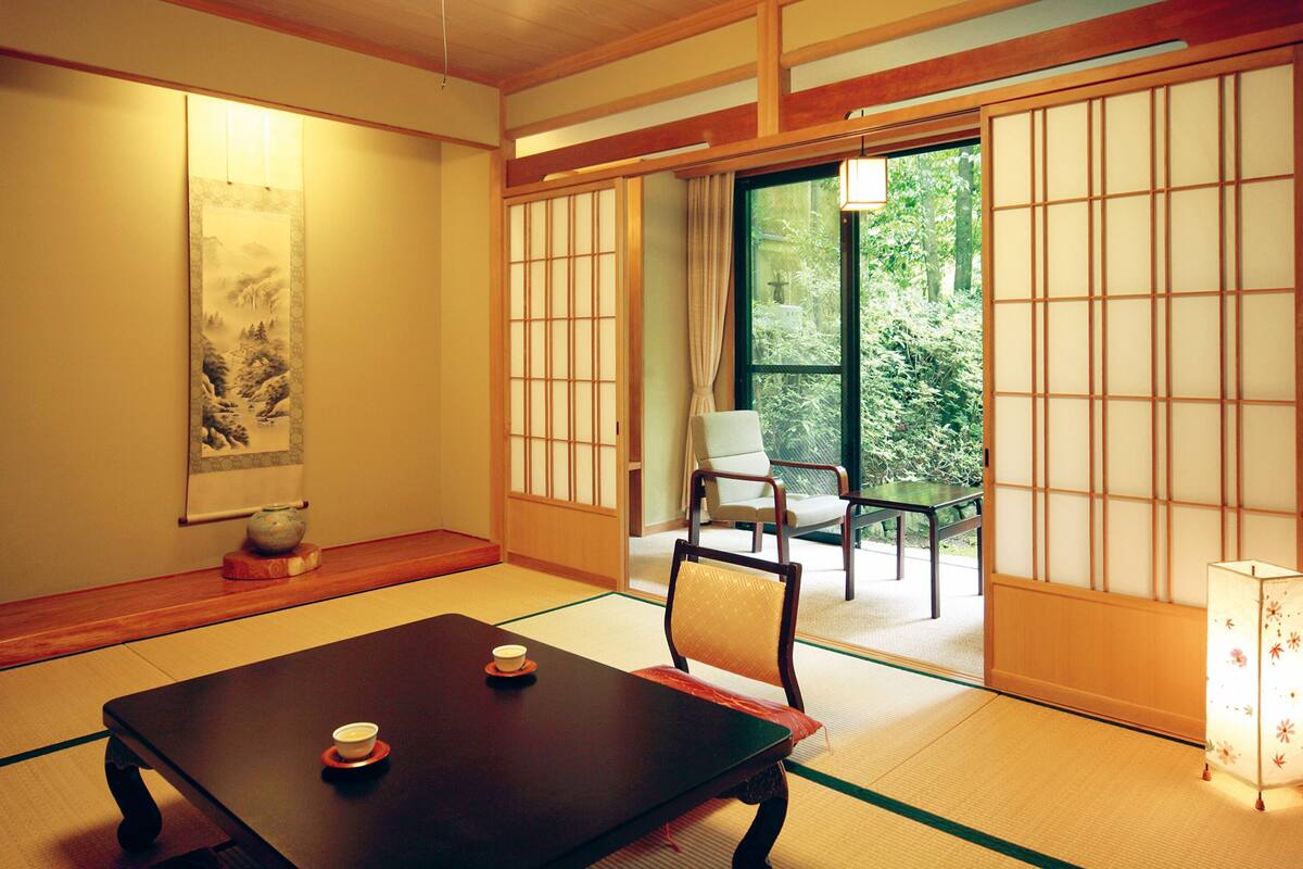 A traditional Japanese room with tatami flooring, a low table with two cups, a chair, and a sliding door leading to a garden.