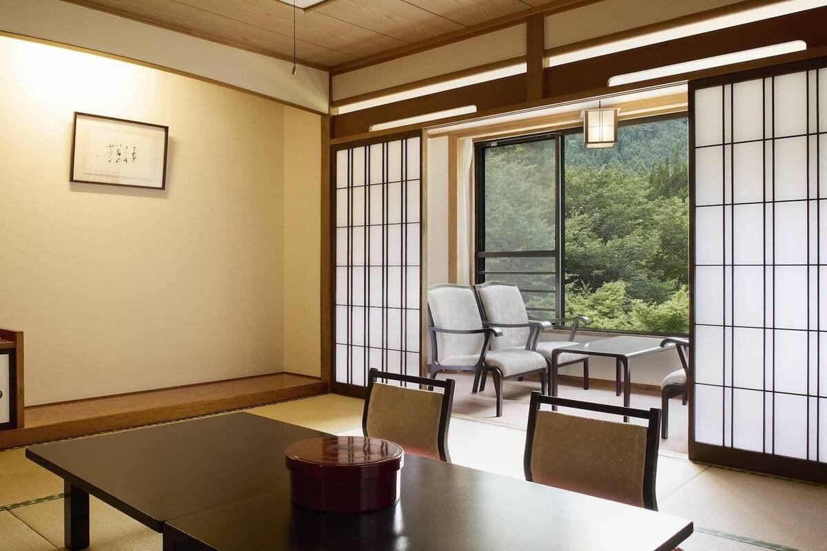 A traditional Japanese room with tatami flooring, a low table, chairs, a shoji screen, and a view of greenery outside.